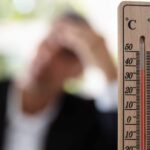 Close-up of a thermometer that reads eighty degrees. A person is in the background pressing their hand to their forehead.