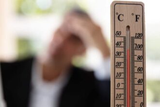 Close-up of a thermometer that reads eighty degrees. A person is in the background pressing their hand to their forehead.