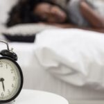 Close-up photo of an alarm clock and a woman sleeping in the background.