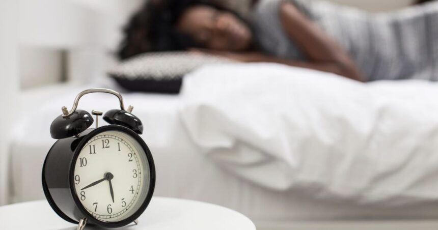 Close-up photo of an alarm clock and a woman sleeping in the background.