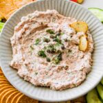 A bowl of smoked salmon dip, garnished with fresh dill and capers, served with crackers and fresh vegetables.