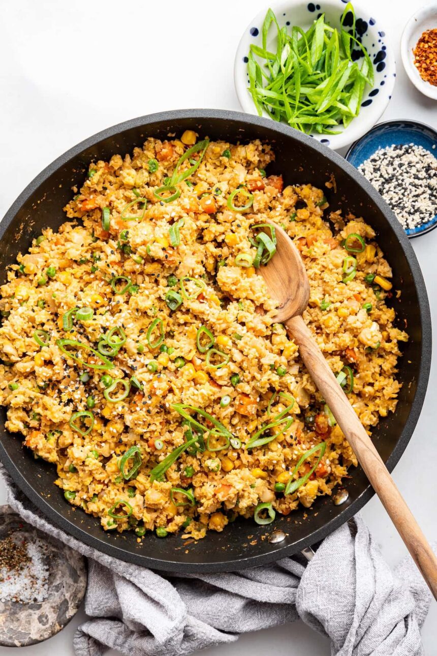 Cauliflower fried rice with eggs in a skillet with a wooden serving spoon. The dish is garnished with fresh green onion.