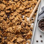 Clusters of chocolate chip cookie granola on a silver baking sheet served with a wooden spoon.