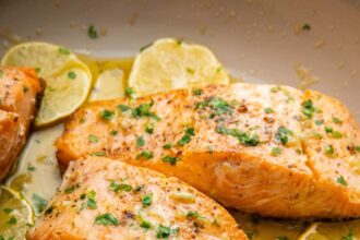 Four salmon filets in a pan with garlic butter and lemon wedges. The filets are garnished with fresh herbs.