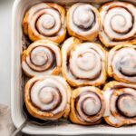 A white square baking dish filled with 9 baked cinnamon rolls that are topped with a glaze.