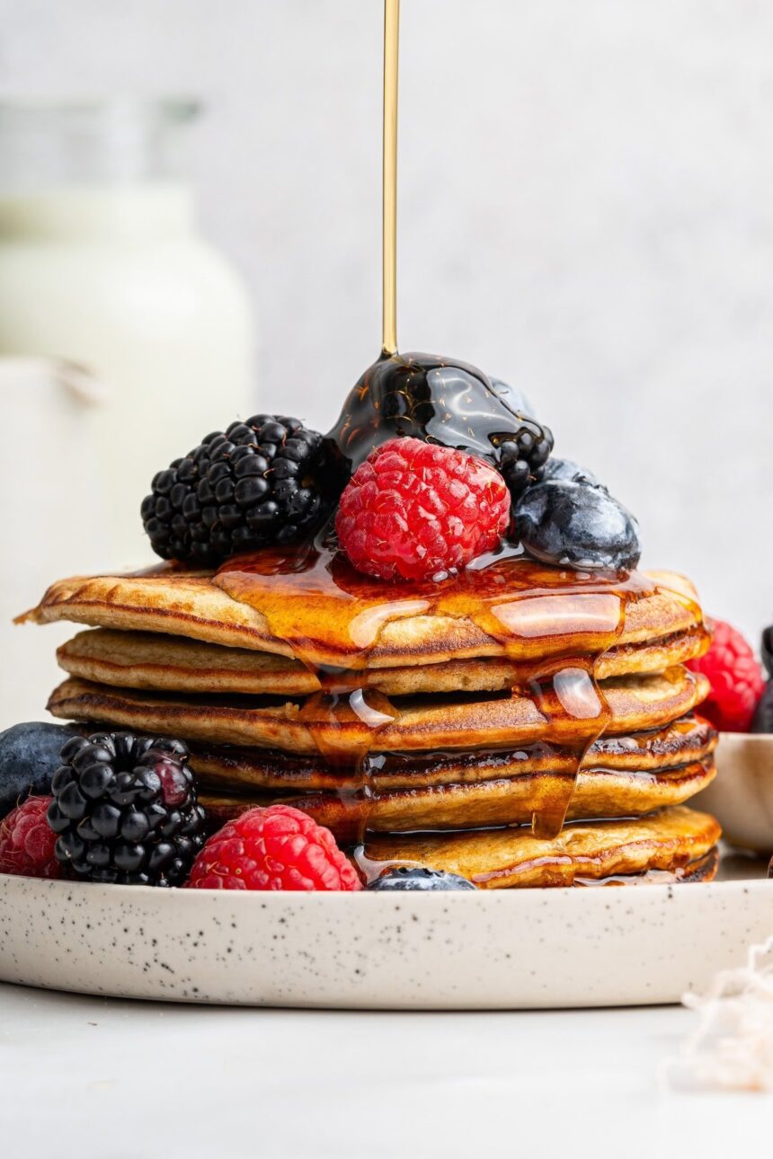 Maple syrup being poured on top of a stack of six greek yogurt pancakes on a plate. They