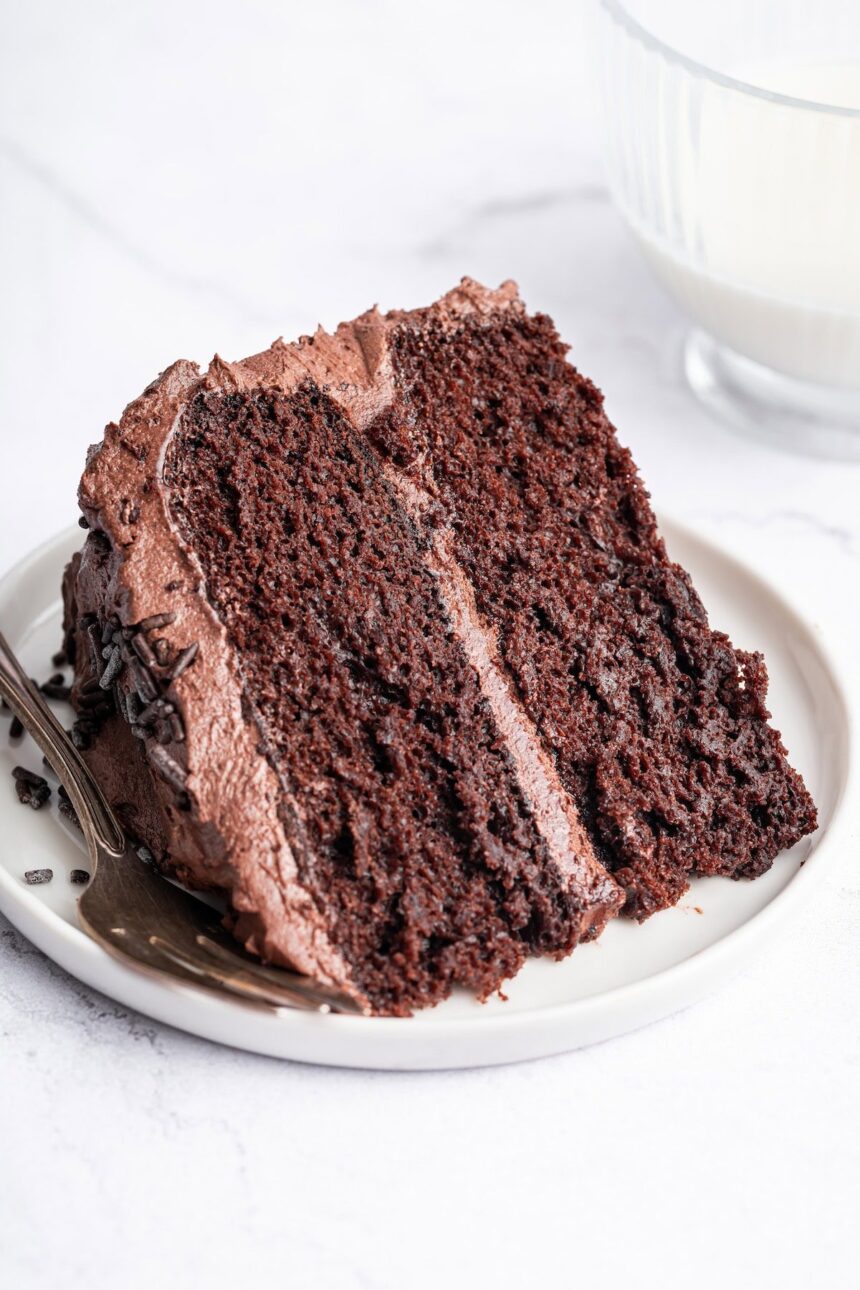 A slice of chocolate cake with chocolate buttercream frosting on a white plate with a metal fork to the side.