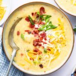 A bowl of potato soup with a silver spoon in it. The soup is topped with bacon bits, green onion, shredded cheese and sour cream.